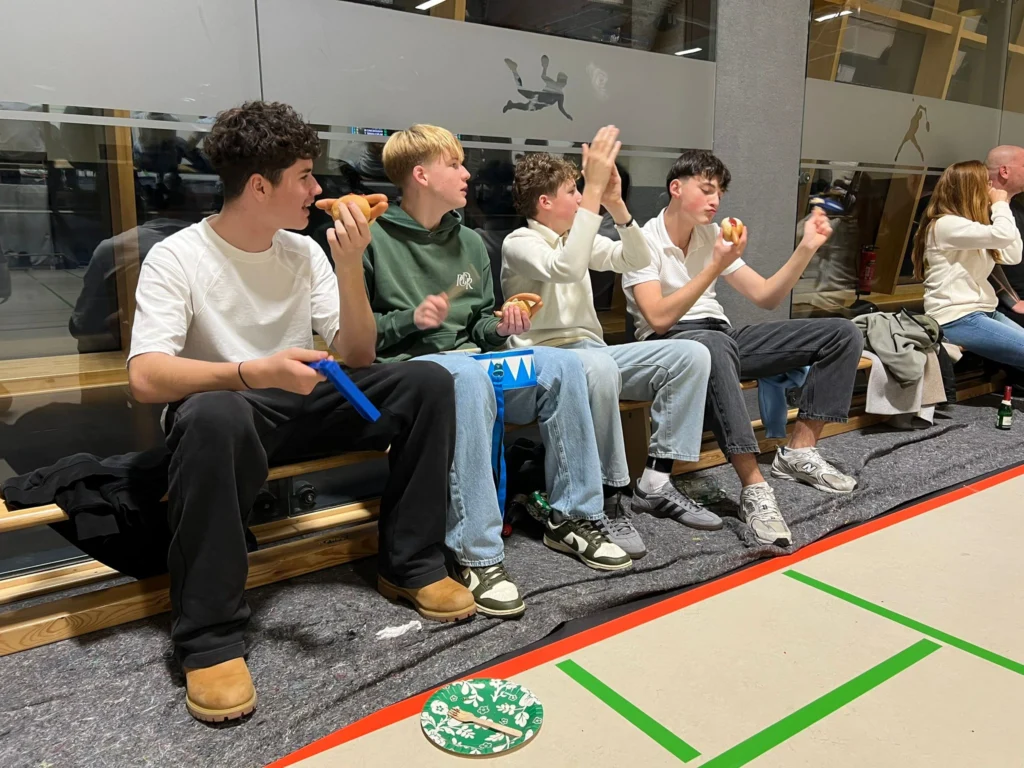 Publikum beim Heimspieltag. 4 allpaudierende Jugendliche, die auf einer Bank sitzen